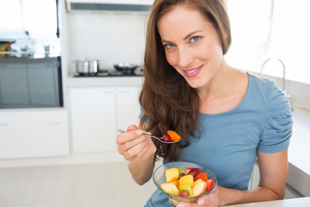 Sorridente jovem comendo salada de frutas na cozinha