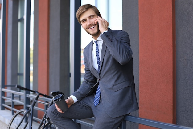 Sorridente jovem elegante usando telefone celular enquanto bebe café ao ar livre com a bicicleta.