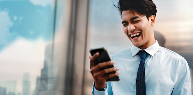 Sorridente jovem empresário usando telefone celular na cidade urbana. O edifício moderno reflete na parede. Visão de baixo ângulo