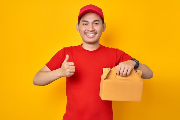 Sorridente jovem entregador asiático em roupa de trabalho de camiseta de boné vermelho trabalha como correio de revendedor segurando sobremesa de bolo em caixa vazia sem marcação, mostrando o polegar para cima gesto isolado no fundo amarelo Conceito de serviço