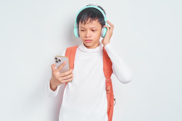 Sorridente jovem estudante asiático com chapéu de natal e roupas casuais com fones de ouvido de mochila para ouvir música via telefone celular isolado no fundo branco