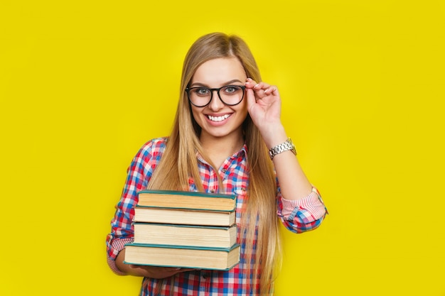 Sorridente jovem estudante elegante está de pé com livros na parede amarela em copos e roupa brilhante casual