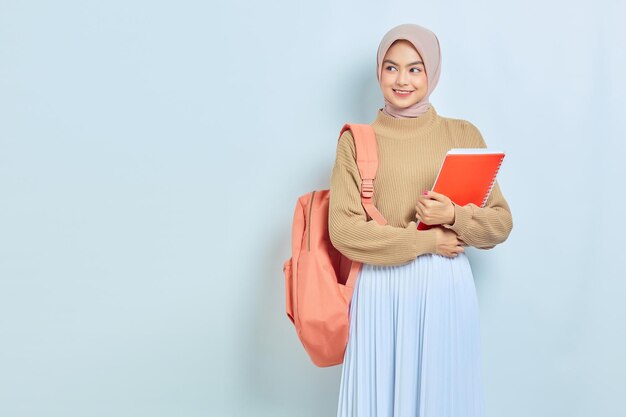 Sorridente jovem estudante muçulmana asiática em suéter marrom e mochila segura livro olhando para o espaço de cópia isolado no fundo branco de volta ao conceito de escola