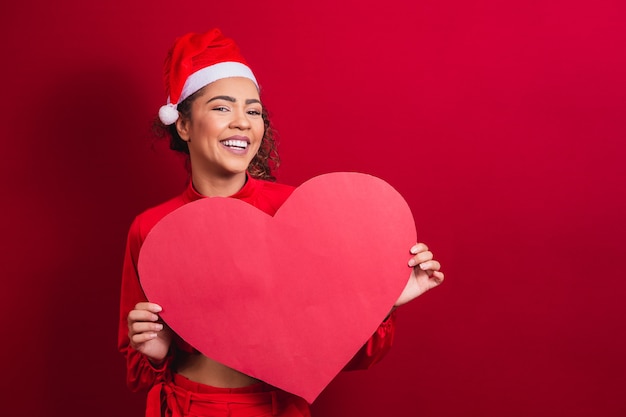 Sorridente jovem garota afro com chapéu de Natal, segurando um cartão de coração enorme com espaço livre para texto.