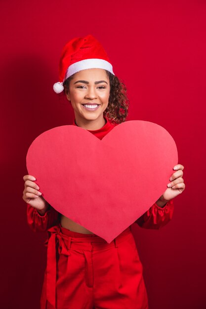 Sorridente jovem garota afro com chapéu de Natal, segurando um cartão de coração enorme com espaço livre para texto.