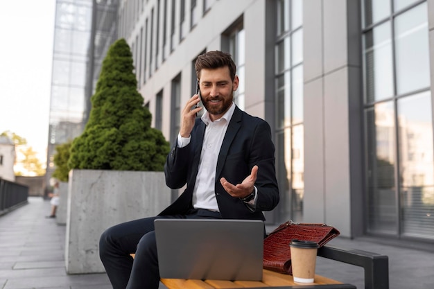 Sorridente jovem gerente europeu ocupado com barba de terno com xícara de café e chamadas de laptop por telefone