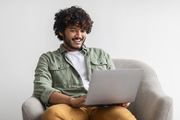 Sorridente jovem indiano sentado na poltrona relaxando com o laptop
