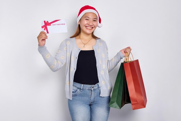 Sorridente jovem loira asiática de 20 anos usando um chapéu de Natal segurando um certificado de vale-presente e uma sacola de compras na mão isolada no fundo branco