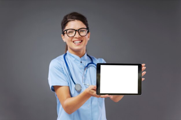 Foto sorridente jovem médica de uniforme, com estetoscópio em volta do pescoço em pé e mostrando o tablet.