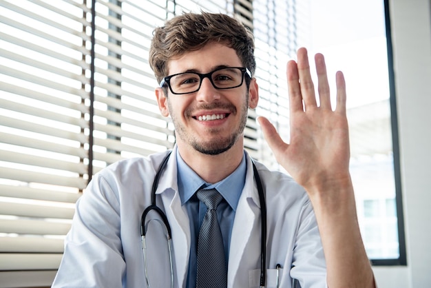 Sorridente jovem médico masculino acenando com a mão para a câmera cumprimentando o paciente on-line via videochamada à distância do escritório