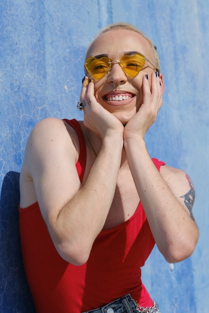 Sorridente jovem modelo não binário usando óculos escuros em forma de coração enquanto posava ao ar livre em um dia ensolarado. Identidade de gênero e conceito não binário.