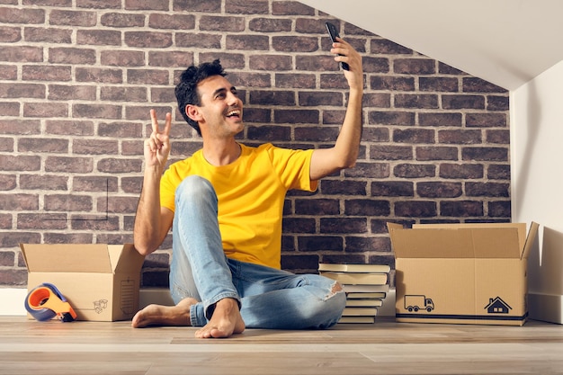 Sorridente jovem moreno com características árabes jeans azul e camiseta amarela homem solteiro que acabou de se mudar para sua nova casa sentado no chão tirando uma selfie