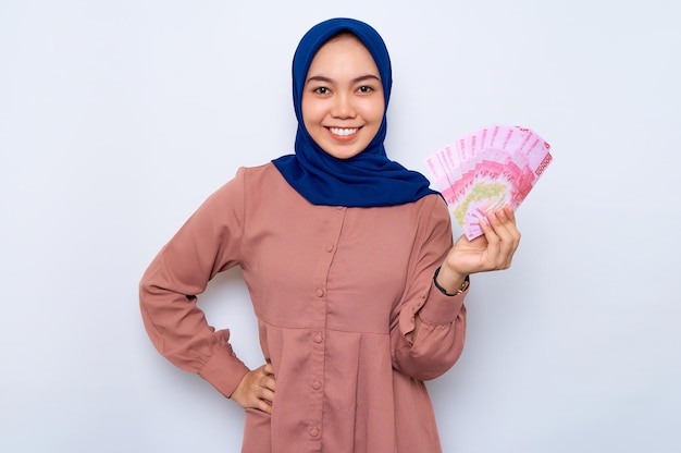 Sorridente jovem muçulmana asiática de camisa rosa segurando notas de dinheiro isoladas sobre fundo branco Conceito de estilo de vida religioso de pessoas