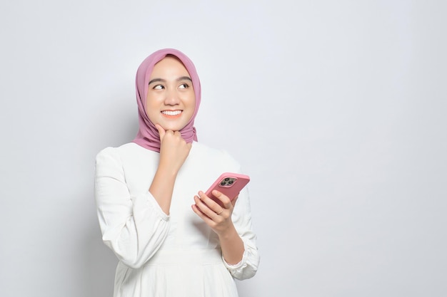 Sorridente jovem muçulmana asiática usando telefone celular e desviando o olhar para o espaço da cópia isolado sobre o fundo branco