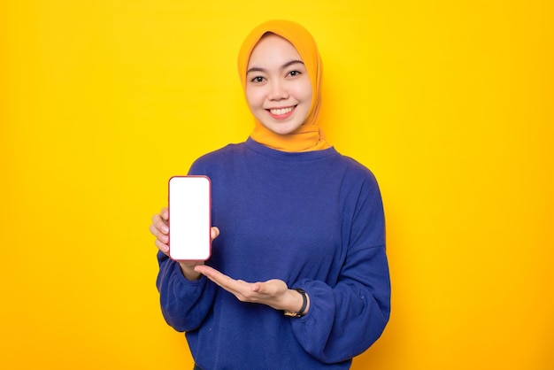 Sorridente jovem muçulmana asiática vestida de suéter casual mostrando celular de tela em branco recomendando aplicativo móvel isolado sobre fundo amarelo