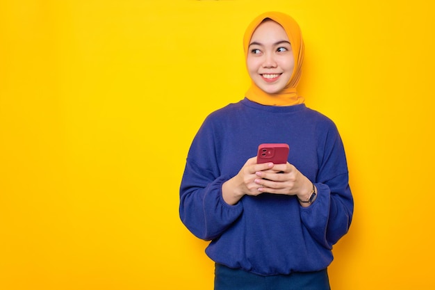 Sorridente jovem muçulmana asiática vestida de suéter casual segurando telefone celular e olhando de lado para o espaço de cópia isolado sobre fundo amarelo