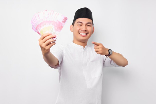 Sorridente jovem muçulmano asiático apontando o dedo para notas de dinheiro em dinheiro rupias isoladas no fundo branco Conceito de estilo de vida islâmico religioso de pessoas