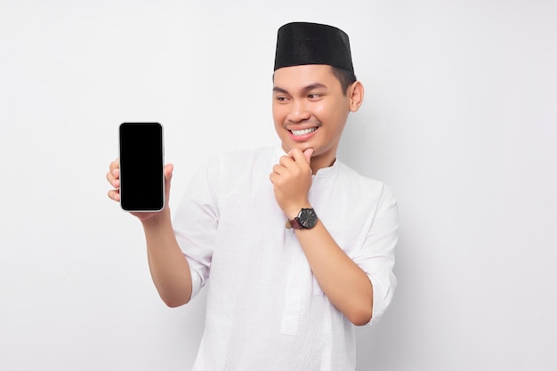 Sorridente jovem muçulmano asiático mostrando a tela do smartphone tocando seu queixo pensando sobre a oferta no fundo branco Celebração do conceito de estilo de vida do Islã religioso do povo Ramadã e Ied Mubarak