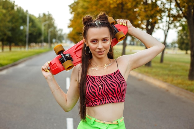 Sorridente jovem patinadora com skate em pé na estrada no verão e olhando para a câmera