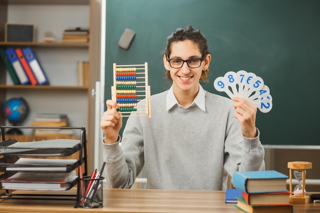 sorridente jovem professor de óculos segurando ábaco com número divertido sentado na mesa com ferramentas escolares em sala de aula