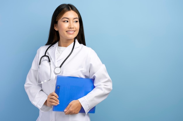 Sorridente médica asiática pediátrica vestindo vestido branco médico segurando a pasta e olhando para longe