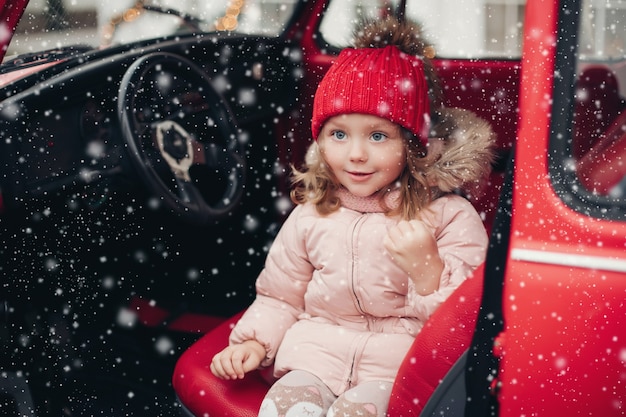 Sorridente menina bonita do inverno com chapéu vermelho, sentado no carro, se divertindo, tiro médio. feliz linda bebê do sexo feminino em roupas quentes, tendo uma emoção positiva ao ar livre, rodeada de flocos de neve, curtindo a infância