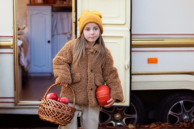 Sorridente menina de chapéu amarelo com cesta de maçãs vermelhas e uma abóbora nas mãos dela.
