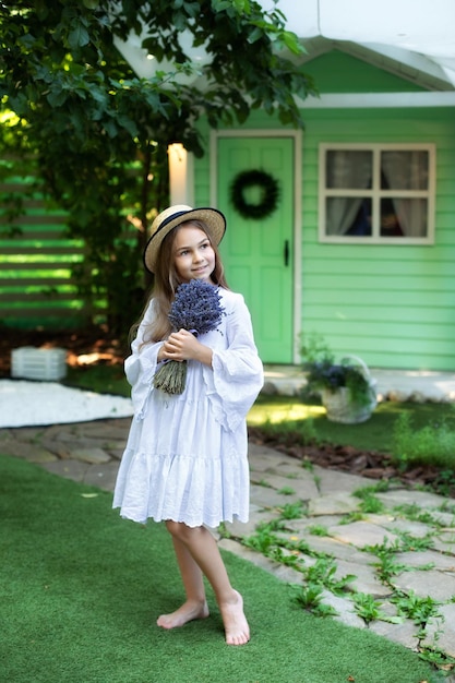 Sorridente menina de vestido branco e chapéu com buquê de lavanda no gramado verde no jardim.