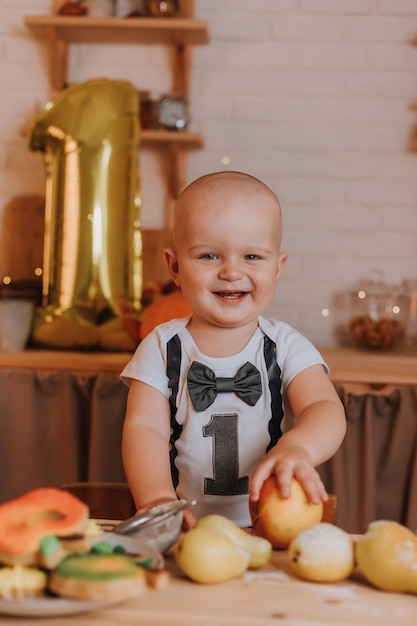 Sorridente menino de um ano de idade em roupas festivas tem uma maçã nas mãos. primeiro aniversário. corpo com o número um. bola de folha dourada. lanche saudável para crianças. pequeno cozinheiro. Foto de alta qualidade