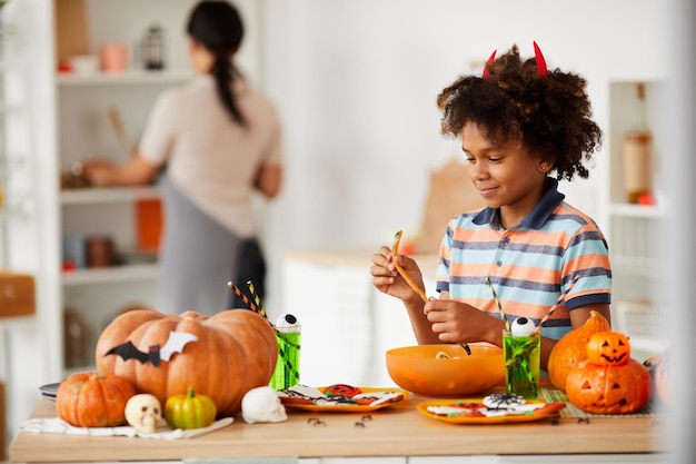 Sorridente menino negro bonito com chifres de demônios em pé no balcão com vários doces de halloween e eati