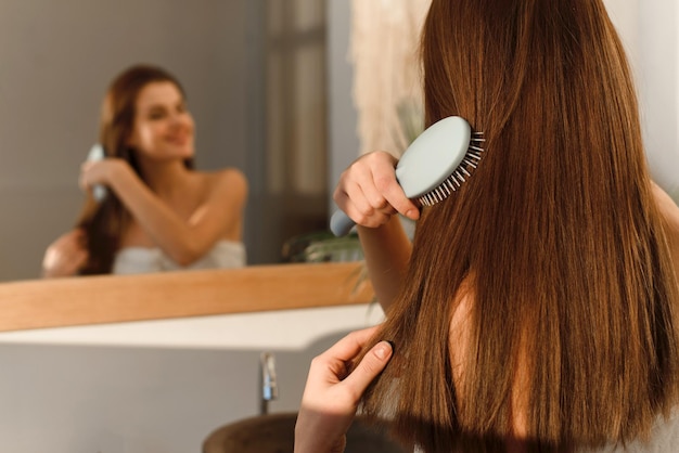 Foto sorridente morena bonita em uma toalha penteia seu cabelo longo e sedoso com um pente de manhã após procedimentos cosméticos no fundo do banheiro