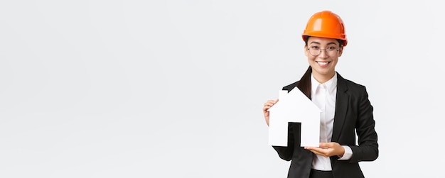 Sorridente vendedora asiática feminina de empresa de construção que vende casas usa capacete e terno de negócios mostrando maket de casa aconchegante em pé fundo branco