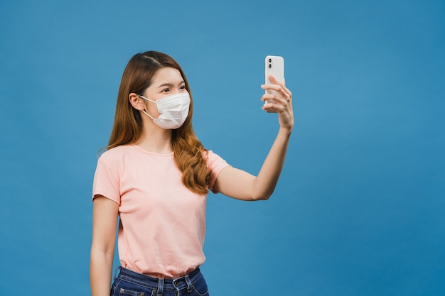 Foto sorrindo adorável mulher asiática usando máscara médica fazendo selfie foto no telefone inteligente com expressão positiva em roupas casuais e suporte isolado na parede azul