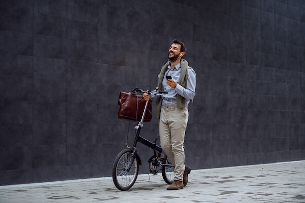 Sorrindo alegre empresário elegante caucasiano, segurando o telefone inteligente, ouvindo música e empurrando sua bicicleta. No fundo é parede cinza.