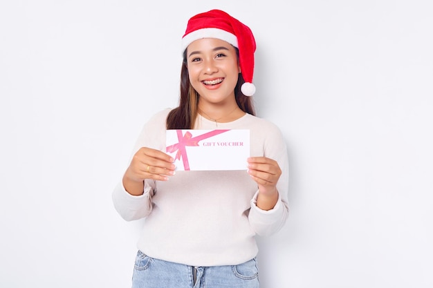 Sorrindo alegre jovem asiática de 20 anos com um chapéu de Papai Noel segurando um cartão de vale-presente para a loja olhando a câmera isolada no fundo branco do estúdio Feliz Natal Conceito