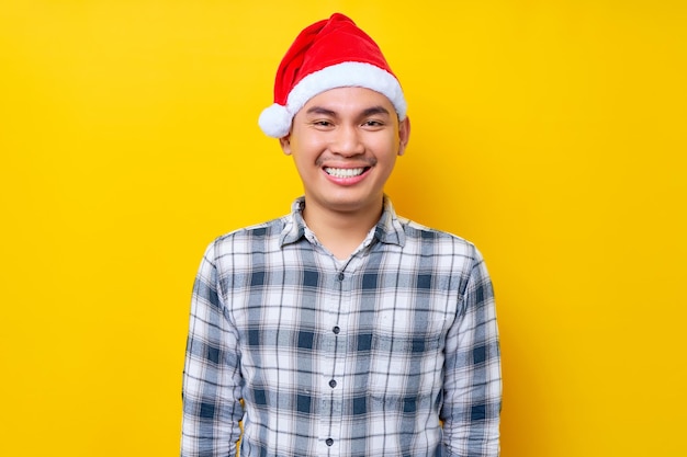 Sorrindo alegre jovem asiático vestindo camisa xadrez no chapéu de Papai Noel olhando para a câmera com expressão confiante na celebração de fundo amarelo estúdio feriado de Natal e conceito de ano novo