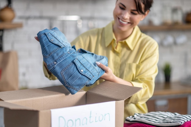 Sorrindo alegre linda jovem colocando um item de roupa dobrado na caixa de papelão