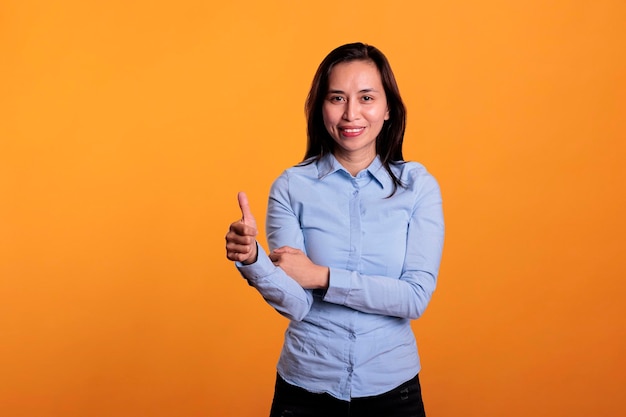 Sorrindo alegre mulher asiática dando polegares para cima gesto, de pé sobre fundo amarelo. Jovem adulto bem sucedido morena mostrando bem e sinal de aprovação no estúdio. Aprovando o conceito de símbolo