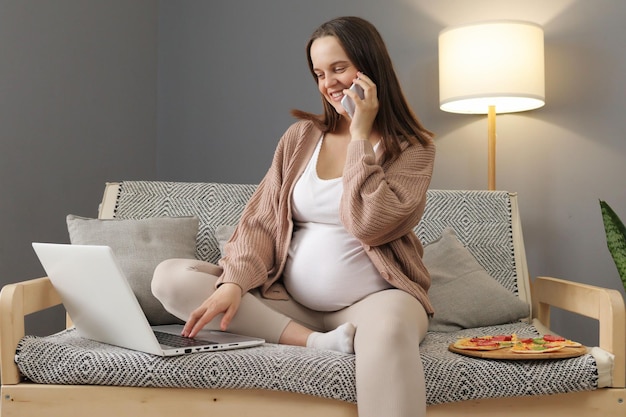 Sorrindo alegremente, satisfeita, grávida freelancer trabalhando em laptop e falando em smartphone com o chefe ou parceiro de negócios comunicando sobre projeto remoto