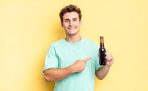 Sorrindo alegremente, sentindo-se feliz e apontando para o lado e para cima, mostrando o objeto no espaço da cópia. conceito de garrafa de cerveja