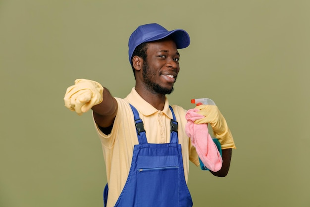 Sorrindo aponta para a câmera segurando o agente de limpeza com pano jovem limpador afro-americano de uniforme com luvas isoladas em fundo verde