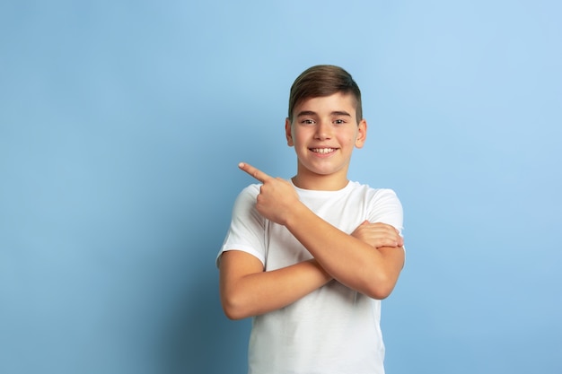 Sorrindo, apontando para o lado. retrato de menino caucasiano isolado no fundo azul do estúdio. lindo modelo masculino adolescente em posar de camisa branca. conceito de emoções humanas, expressão facial, vendas, anúncio.