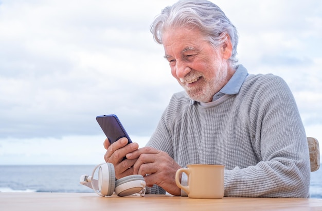 Sorrindo atraente homem adulto sênior maduro com barba usando smartphone ao ar livre no mar