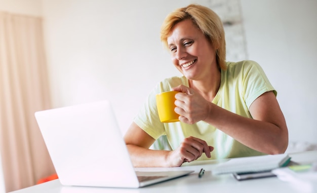 Foto sorrindo bela mulher madura em roupas casuais está relaxando e vendo algum filme de comédia no laptop em casa ou tendo bate-papo on-line com família ou amigos mulher bebe café ou chá