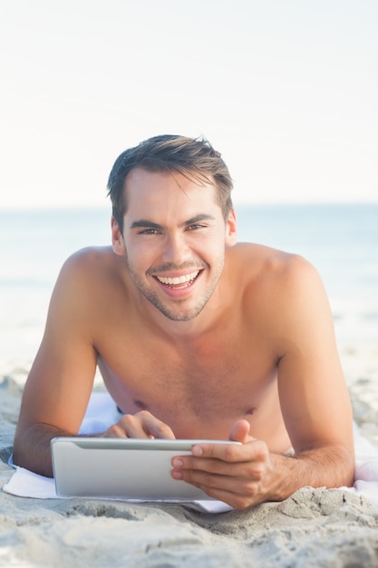 Sorrindo, bonito, deitado na sua toalha, usando seu tablet