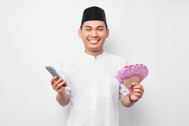 Sorrindo bonito homem muçulmano asiático usando telefone celular e segurando notas de dinheiro em dinheiro rupiah isoladas no fundo branco Conceito de estilo de vida islâmico religioso de pessoas