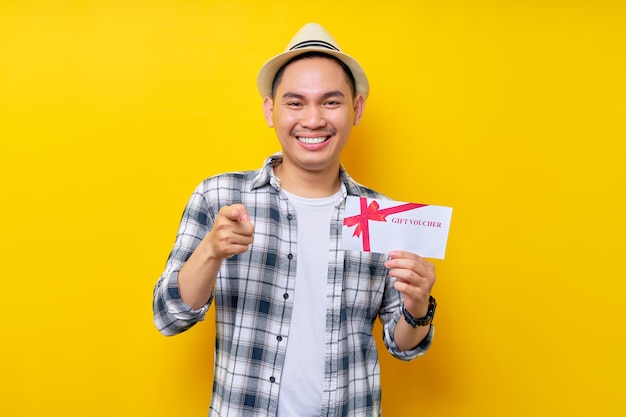 Sorrindo bonito jovem de etnia asiática 20 anos vestindo roupas casuais chapéu segurando vale-presente cupom vale e apontando para você isolado no fundo amarelo Pessoas conceito de estilo de vida