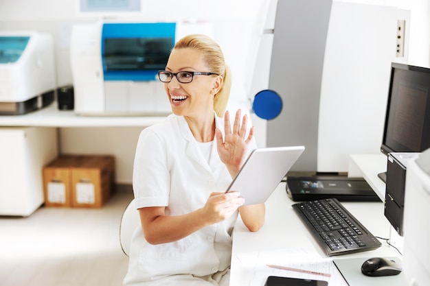 Sorrindo bonito loiro feminino assistente de laboratório em uniforme branco estéril sentado no laboratório e usando o tablet para verificar os resultados da pesquisa.