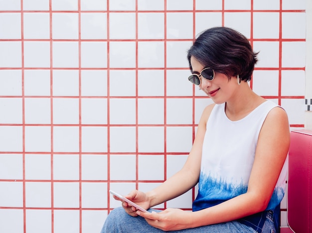 Sorrindo cabelo curto atraente elegante mulher asiática vestindo camisa branca e óculos escuros, sentado no banco vermelho e usando o smartphone no fundo da parede de azulejos brancos e vermelhos com espaço de cópia.