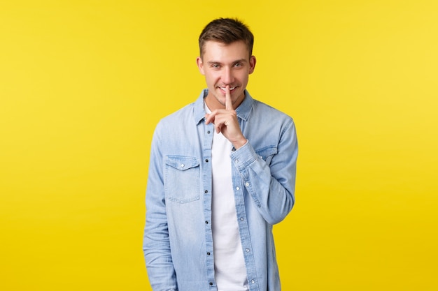 Sorrindo, cara bonito misterioso, mantendo segredo, sorrindo e silenciando a câmera com o dedo indicador pressionado nos lábios, escondendo informações, pedindo para ficar quieto, fundo amarelo de pé.